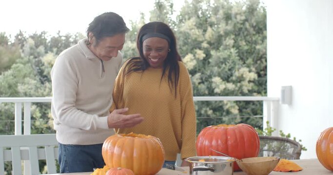 Diverse couple sharing affectionate moment then scooping and carving pumpkins on porch for holiday