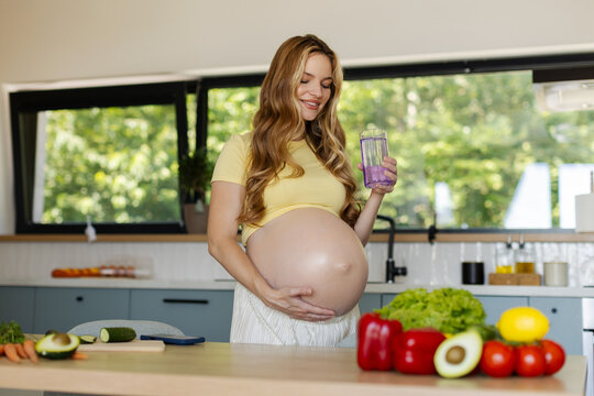 Young pregnant woman drinking glass of water while gently cradling her baby bump and smiling, standing in bright modern kitchen