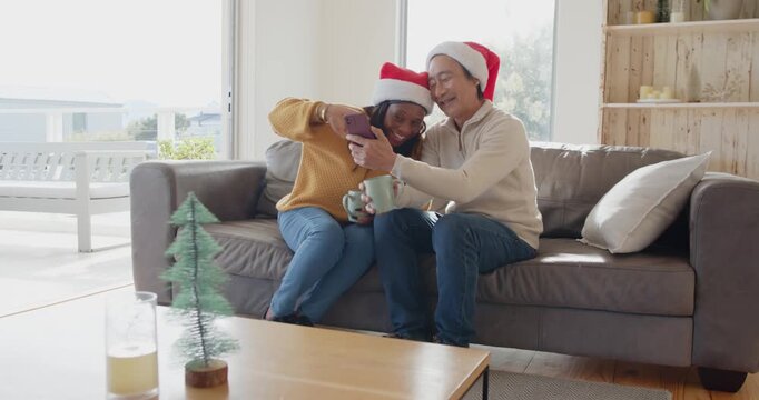 Diverse couple in Santa hats sitting on gray sofa, sharing smartphone during holiday call with mugs