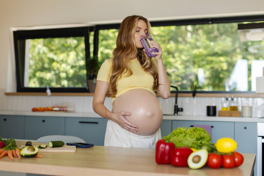 Pregnant woman taking refreshing sip of water while lovingly holding her baby bump in bright and stylish kitchen, copy space