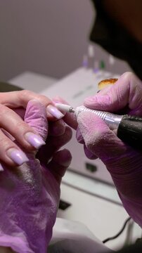 A vertical slow motion close-up video showing a manicurist using an electric drill cutter to remove old coat and shape a client's fingernail in a beauty salon.