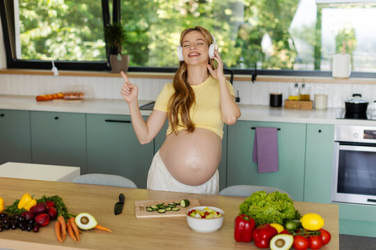 Smiling expectant mother enjoying favorite music through headphones in bright kitchen while preparing homemade salad