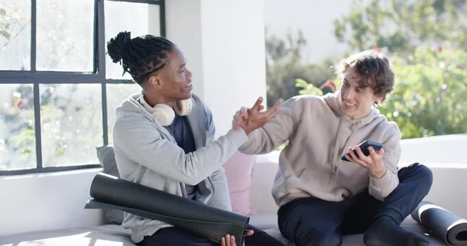 Diverse male friends laughing, high-fiving as right man showing phone on sunlit couch with mat