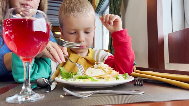 A curious young boy enjoys eating his delicious salad with a fork. He dines happily inside a cozy cruise ship restaurant beside a bright red drink.