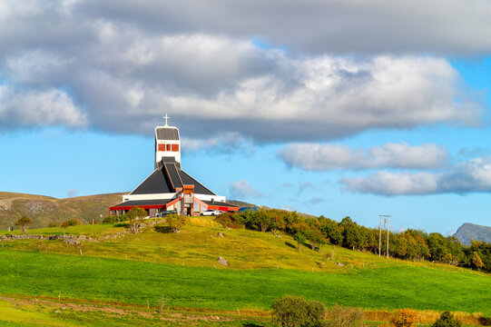 Borge Church in Borg in the Lofoten Islands, standing on a hill surrounded by green meadows and autumn vegetation beneath a blue sky with dramatic clouds.
