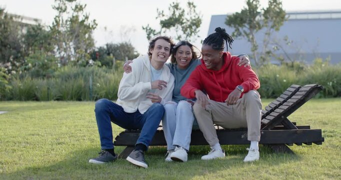 Diverse friends sitting on low bench on lawn, center wrapping arms, laughing and bonding