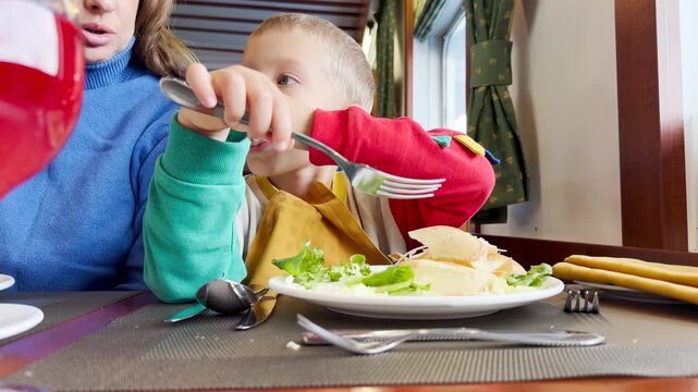 A playful boy holds a fork while grabbing food from his plate. He enjoys a happy mealtime inside a cozy cruise ship restaurant.