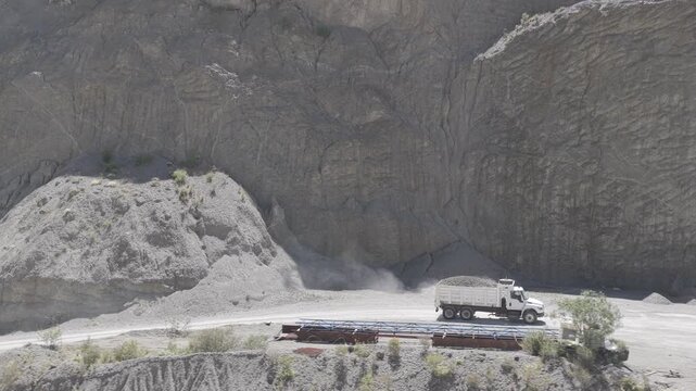 Aerial view of a loaded dump truck driving along a dusty quarry road beneath a towering rock wall while the camera tracks alongside from left to right, D-Log M