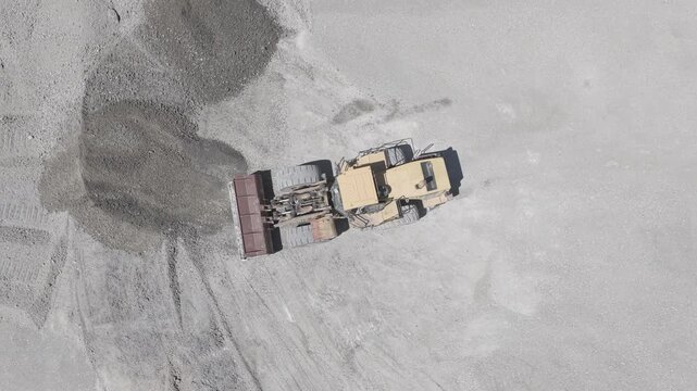 Top-down aerial view of a wheel loader pushing gravel across a sand and gravel quarry while the camera tracks the machine from above, D-Log M