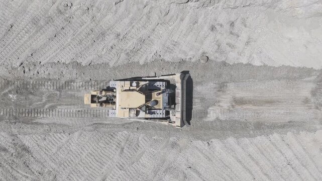 Top-down aerial view of a crawler bulldozer with a rear ripper reversing across a sand and gravel quarry while the camera hovers overhead, D-Log M