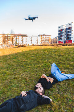 Two young men lying on grass in an urban park watching a drone flying in the sky