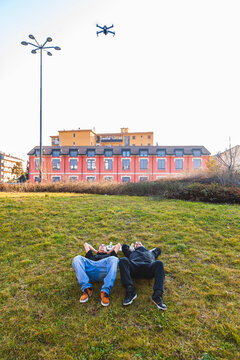 Two young men lying on grass observing a drone flying in the sky