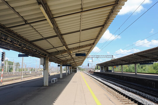 Empty railway station platform. Metal canopy structure. Parallel train tracks background. Overhead electric railway lines. Sunny blue summer sky. Modern transport infrastructure. Quiet travel setting.
