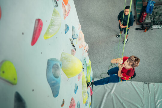 High-angle POV of a young boy climbing a colorful indoor artificial rock wall while his mother carefully belays him from the gym floor below during their active family training and sports session.