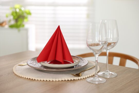 Red napkin folded in shape of pyramid, plates, cutlery and glasses on wooden table indoors