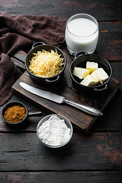 Grated yellow cheese in a black ramekin beside solid butter cubes and powdered flour in glass bowls, a glass of milk and brown spice on a dark wooden cutting board with a metal knife
