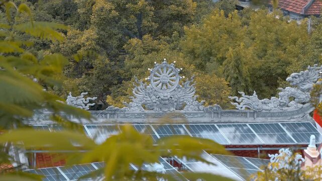An intricately carved stone Dharmachakra or Wheel of Dharma held by dragons rises above lush green foliage of the park and a bunch of racks with solar panels in the Long Son Pagoda in Nha Trang, Vietn