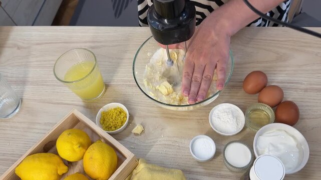 A collection of baking ingredients and tools arranged on a light wooden table surface, showing hands actively mixing ingredients in a bowl with an electric mixer
