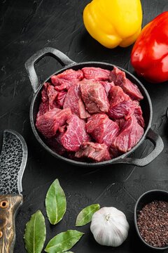 Raw diced beef chunks in a cast iron frying pan with red and yellow bell peppers, whole garlic bulb, bay leaves, and peppercorns on black stone surface under directional light
