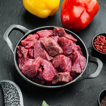Diced raw beef chunks in a cast iron skillet beside red and yellow bell peppers on a dark textured stone surface with peppercorns in a small bowl under directional lighting