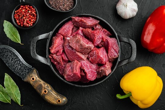 Raw lean beef chunks in a cast iron frying pan surrounded by red and yellow bell peppers garlic whole peppercorns and bay leaves on a black textured stone surface under even overhead light