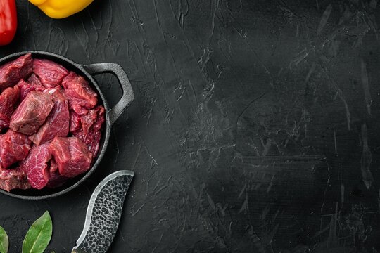 Black textured stone surface with diced raw beef chunks in cast iron frying pan beside sharp knife and fresh bay leaves, red and yellow bell peppers partially visible