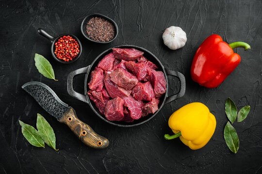 Red and yellow sweet bell peppers beside diced raw beef chunks in a cast iron frying pan with whole garlic bulb, bay leaves, black and red peppercorns