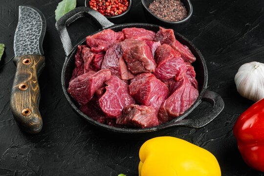 Raw chopped lean beef chunks in a cast iron frying pan beside red and yellow sweet bell peppers, whole garlic bulb, small bowls of black and red peppercorns, bay leaves