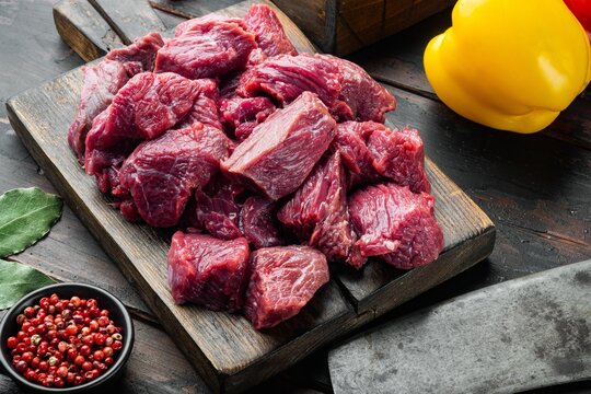 Diced raw chuck beef arranged on a weathered dark wooden cutting board beside whole yellow bell pepper and small black bowl of red peppercorns, fresh bay leaves scattered nearby