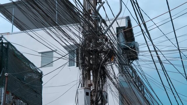A heavy, tangled mass of electrical power cables and communication wires chaotically clustered tightly in a web around a utility pole in front of a white building. Shot in motion
