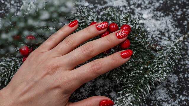 Elegant hand featuring festive red and white Christmas nail art, surrounded by snowy winter scenery, evergreen branches, and bright red berries, creating a cozy holiday atmosphere.