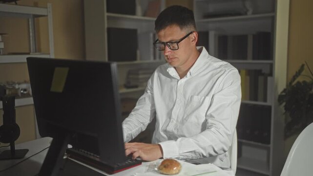 Man holding sandwich at computer, hands unwrapping snack while glancing at watch in building; night fatigue.