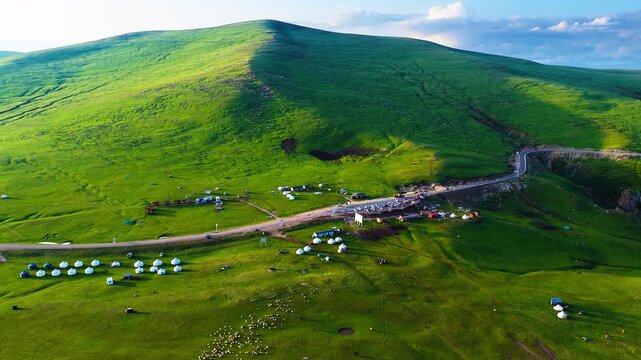 High angle view of scenic grassland mountain with yurts settlements and winding road during sunny day in Xinjiang, China.