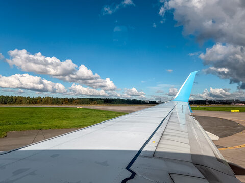 Passenger plane airplane wing view from a window with a blue upward winglet tip on an airport runway ground. Beautiful clear summer sky background template with dramatic white cumulus clouds. 