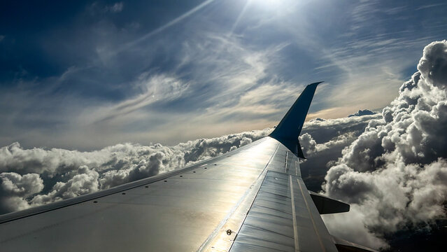 Passenger plane airplane wing view from a window flying high above dramatic white cumulus clouds. Beautiful deep blue sky sky background with bright sun beams rays flare, sunburst, unique cloudscape. 