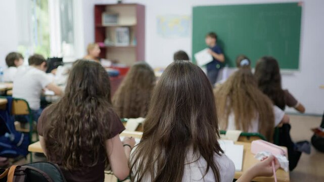 Students sitting at desks in a classroom while a classmate presents at the green chalkboard