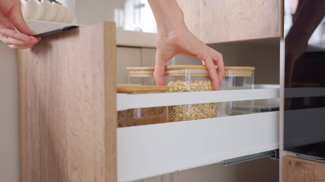 Woman opens pull out pantry drawer and removes clear container with oats from kitchen storage. Concept of food organization, pantry access and home routine