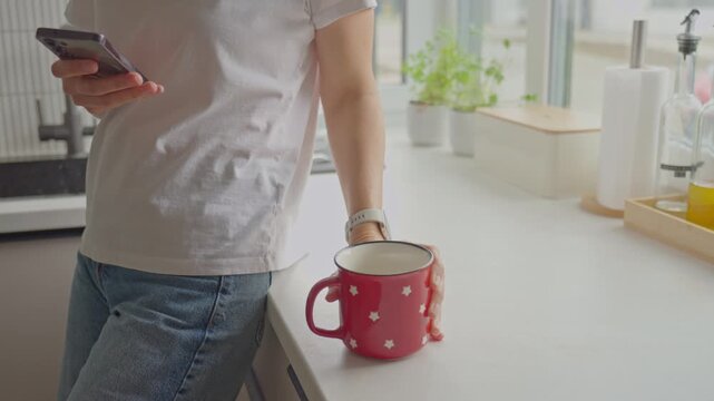 Woman checks smartphone while lifting red mug and taking a drink in kitchen. Concept of mobile technology, everyday routine and multitasking at home