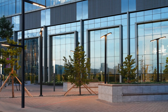 Modern commercial building exterior with glass facade, street lights, and young trees at dusk. Real Photo