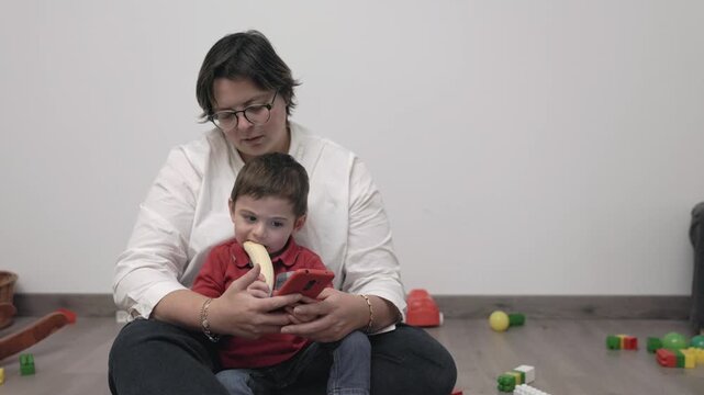 Young boy eating banana watching videos on smartphone with his mother