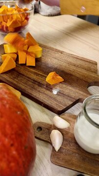 a person cuts orange squash into cubes on a wooden board, pieces of raw garlic and a jar of white liquid sit nearby on a light wooden table