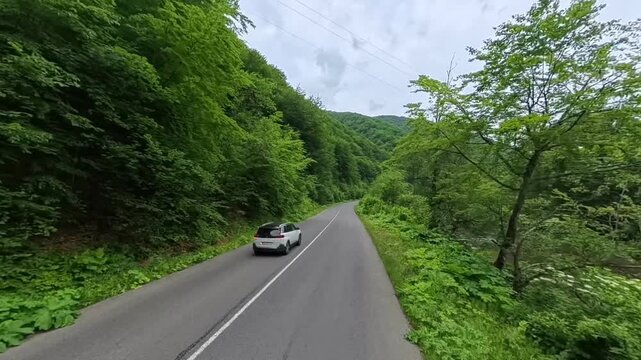 First-person POV driving shot on an asphalt mountain road passing lush green forest hills with a oncoming white car and dramatic cloud formations overhead.