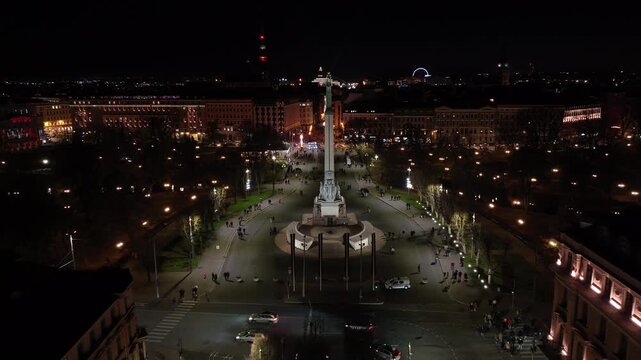 Night descent to Freedom Monument Milda golden stars from drone, Riga. Aerial shot lowering past lit facades, park lights and crowds of Riga, Latvia.