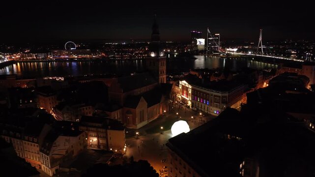 Riga old town at night to St Peters tower beacon from drone, Latvia. Aerial shot passing Dome cathedral, glowing streets, lit bridges and National Library of Riga, Latvia.