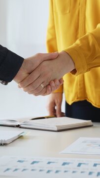 Business partners shaking hands after successful meeting in office while another person is clapping hands over financial documents, laptop and notebook
