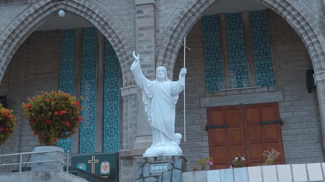 A white marble sculpture of Jesus Christ holding a processional cross outside the Christ the King Cathedral (also known as the Stone Church or Nha tho Nui) in Nha Trang, Vietnam. Set against the stone