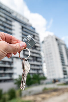 Hand Holding House Keys with House Shaped Keychain against New Apartment Complex Vertical