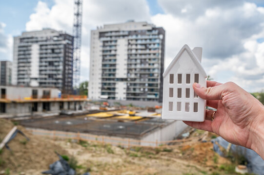 Hand Holding Ceramic House Model with Modern Building Construction and Crane Background Concept