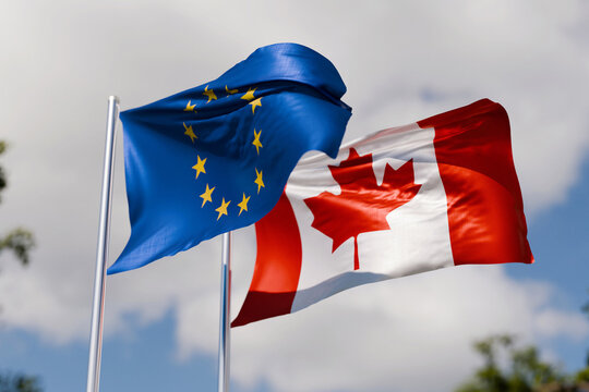 Waving official flags of the European Union (EU) and Canada on metallic poles on a background of blue sky and light clouds. Represents international diplomatic relations, trade, cooperation.