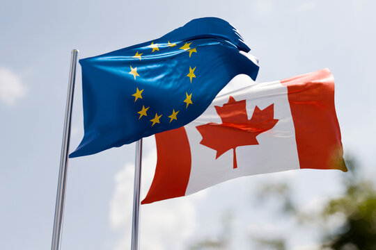 Flags of the European Union and Canada waving in the wind against a blue sky, symbolizing international relations, diplomatic alliance, economic partnership, trade agreements, and cooperation.